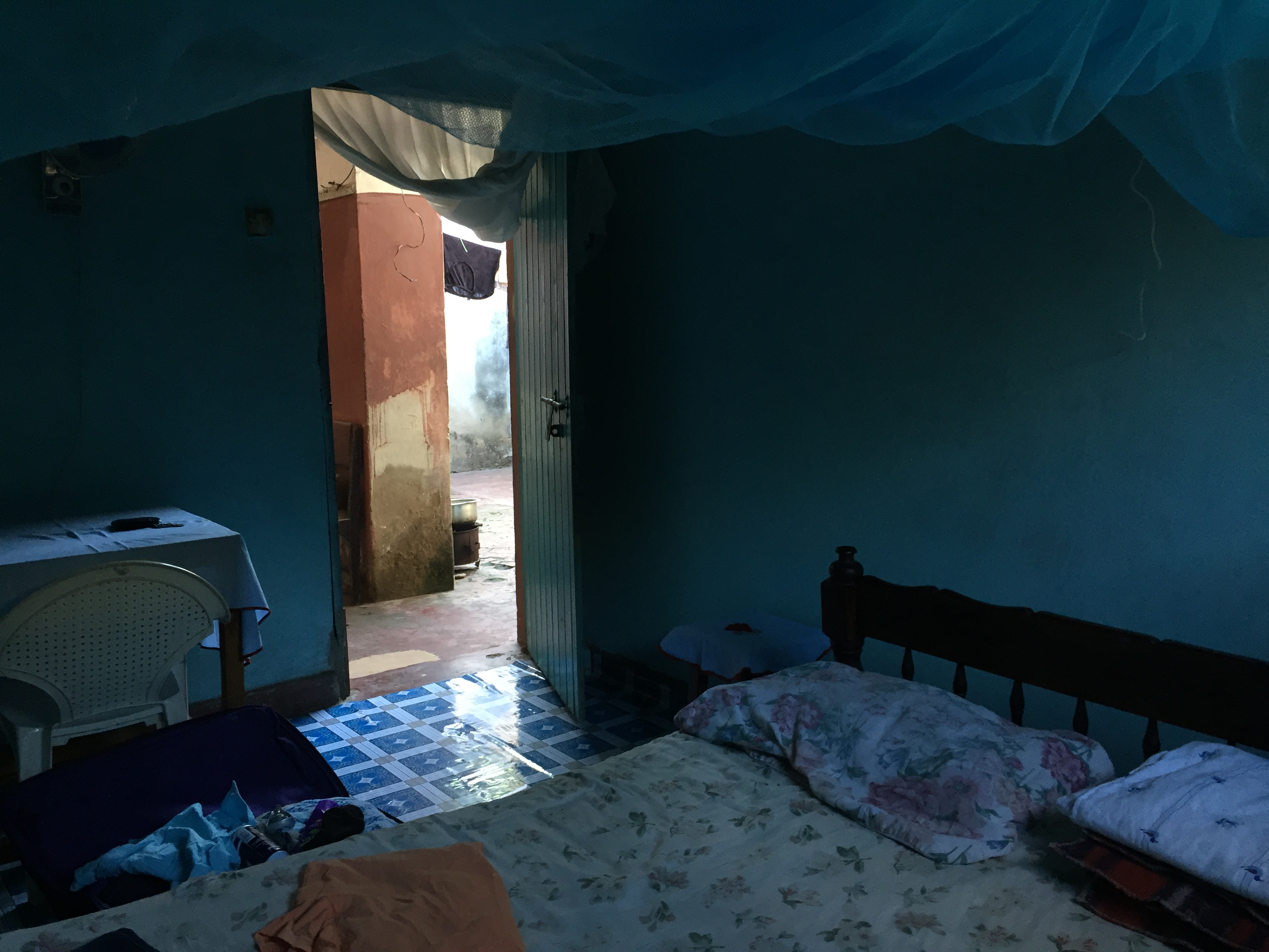 Dimly lit bedroom painted blue, showing bed with mosquito net hanging overtop and an open doorway to red concrete patio